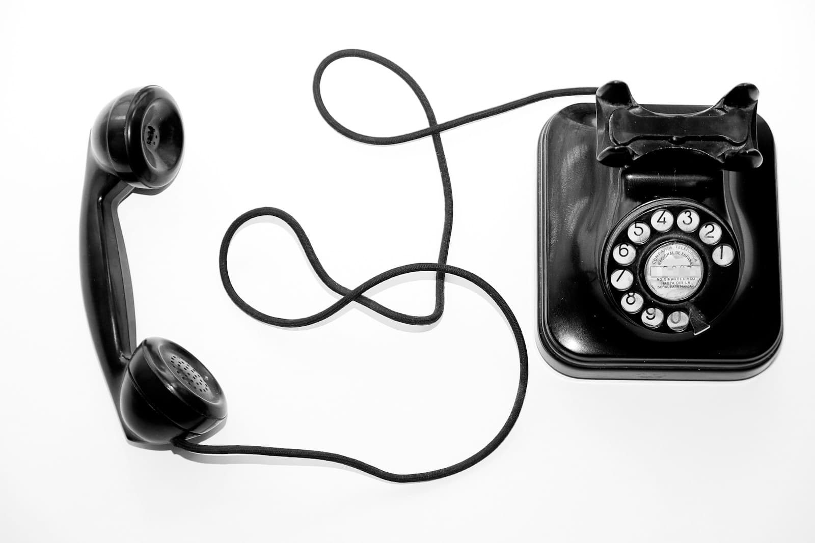Black and white photograph of a vintage rotary telephone with its handset off the hook, the cord looping across a white surface.