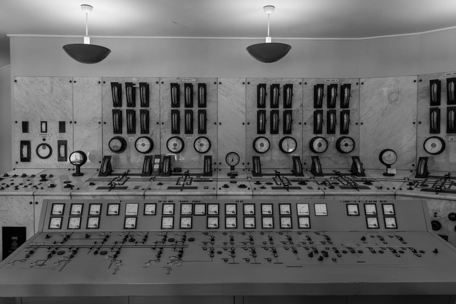 Black and white photograph of a power-station control room wall lined with analog gauges and dials above a switch console.